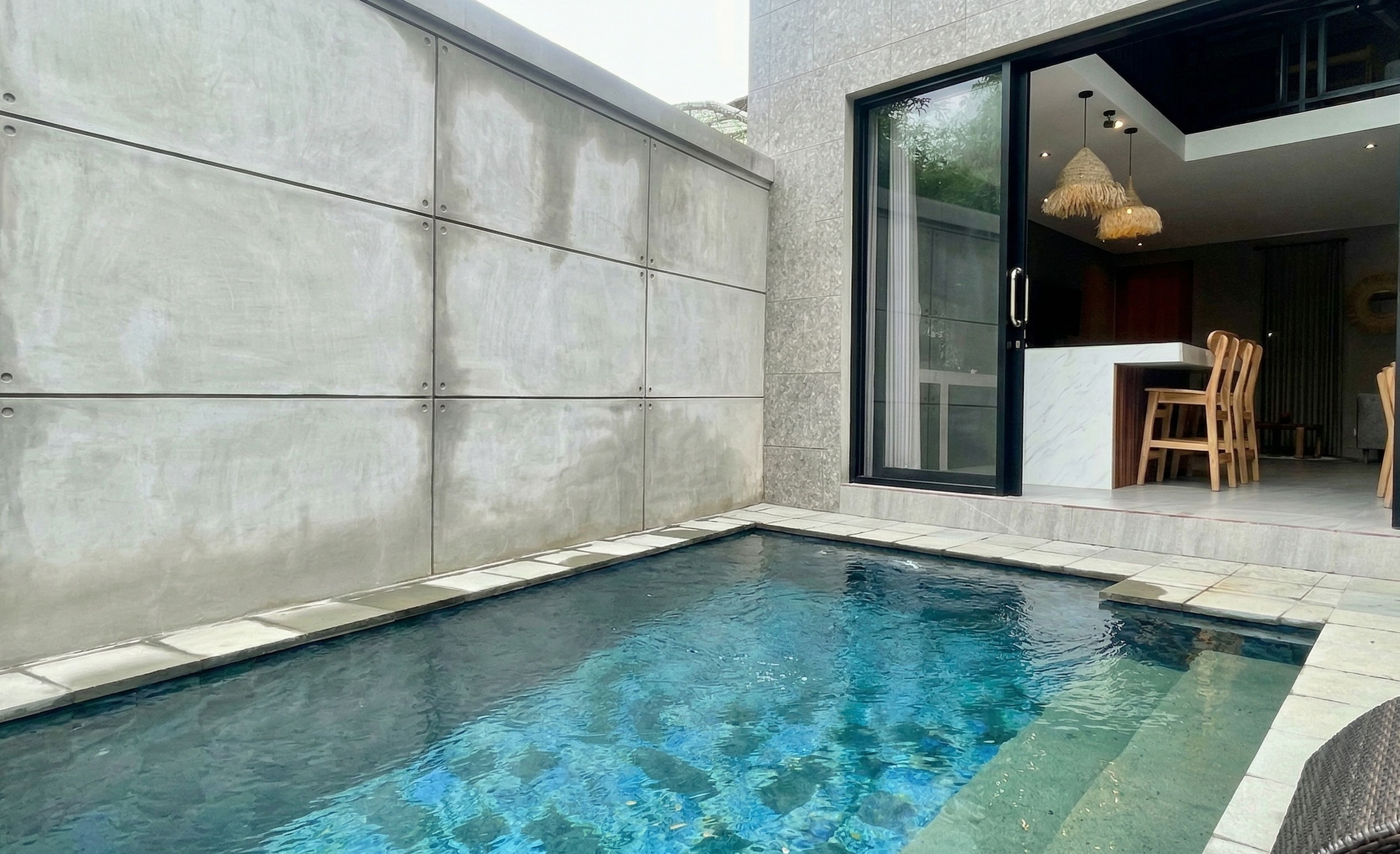 A swimming pool with a modern design, a dining area with wooden chairs, and a glass door leading to a kitchen.
