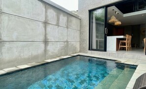 A swimming pool with a modern design, a dining area with wooden chairs, and a glass door leading to a kitchen. - Juminten Villa  (Ubud)