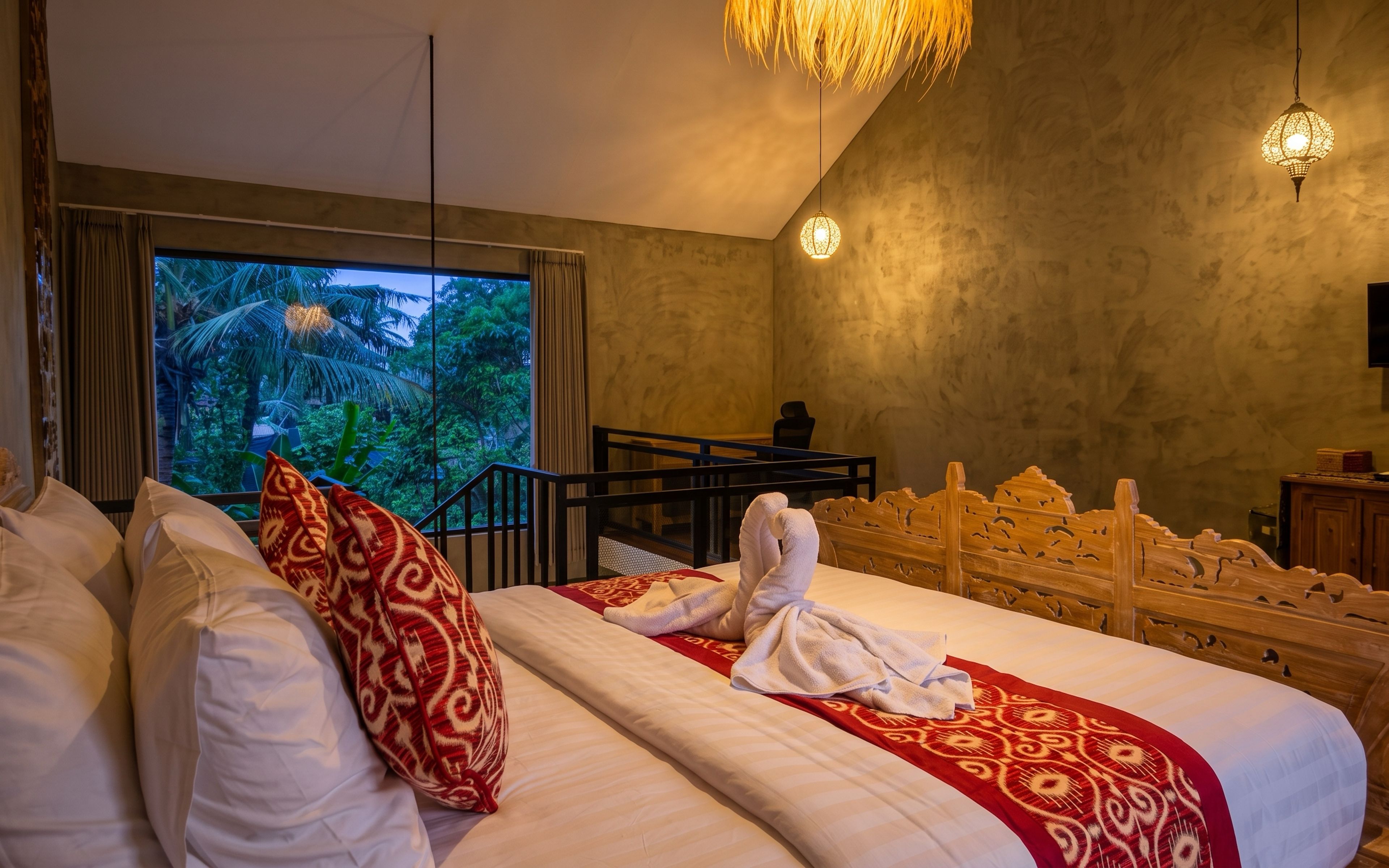 A hotel room with a bed, pillows, a view of greenery, a wooden headboard, and a hanging light fixture.