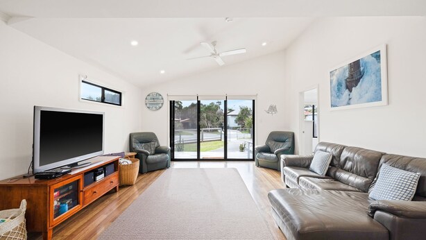 A spacious living room with a flat-screen TV, leather sofas, a wooden cabinet, and a large window offering a view of the outdoors.