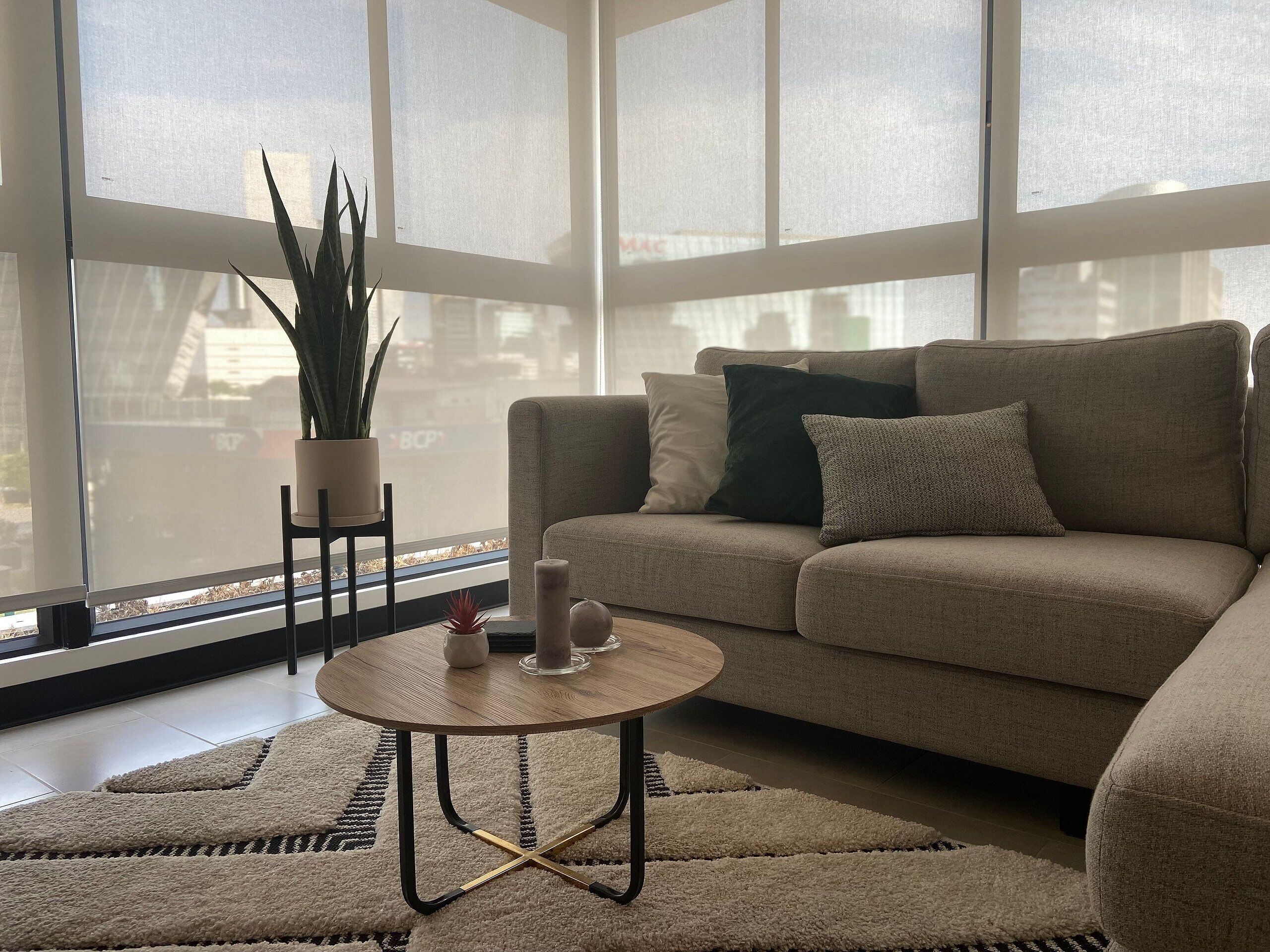 Een woonkamer met een beige bank, een ronde salontafel, een plant in pot en uitzicht door grote ramen.