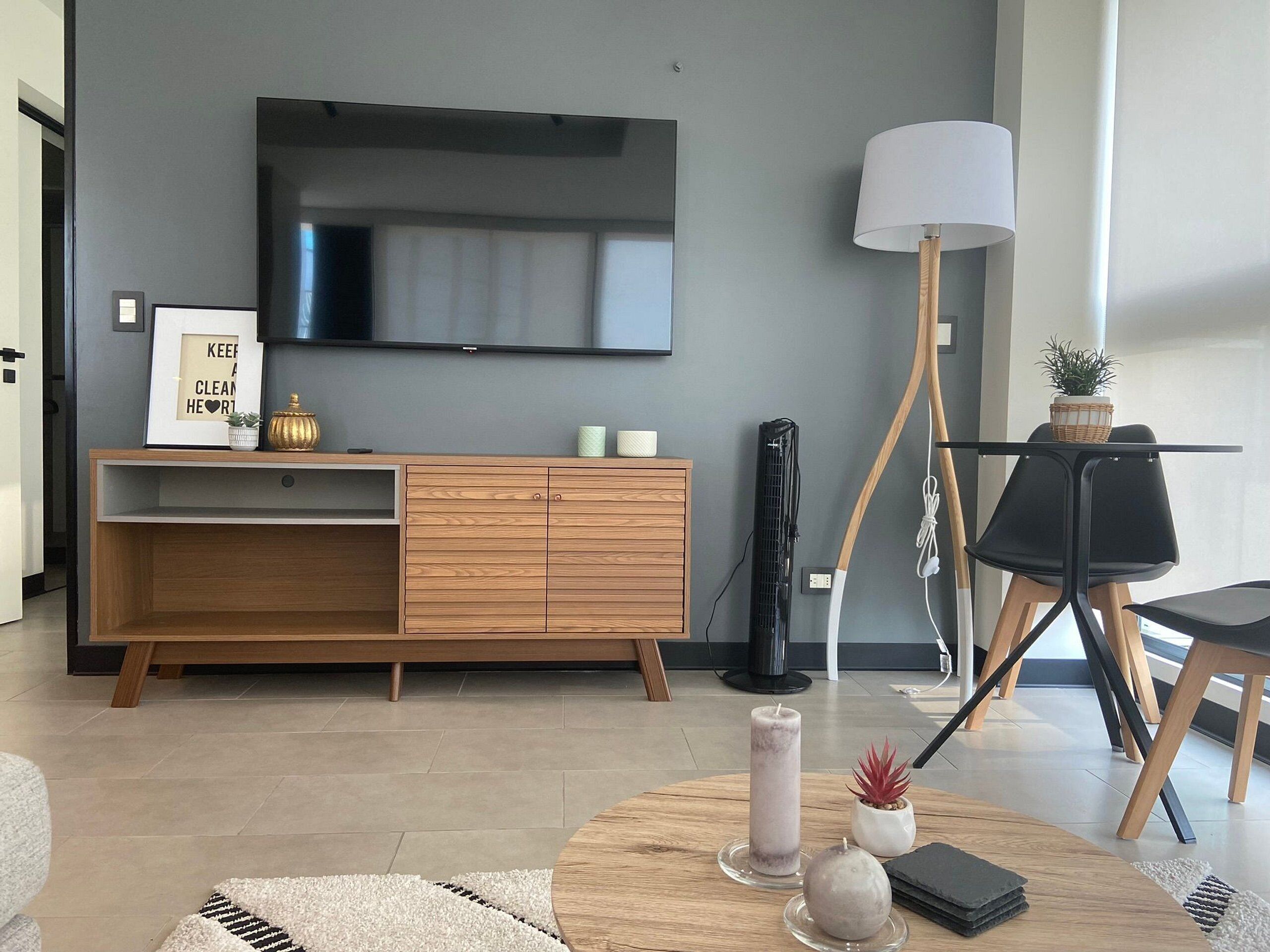 A modern living room with a wooden cabinet, a flat-screen TV, a standing lamp, and a round coffee table with decorative items.