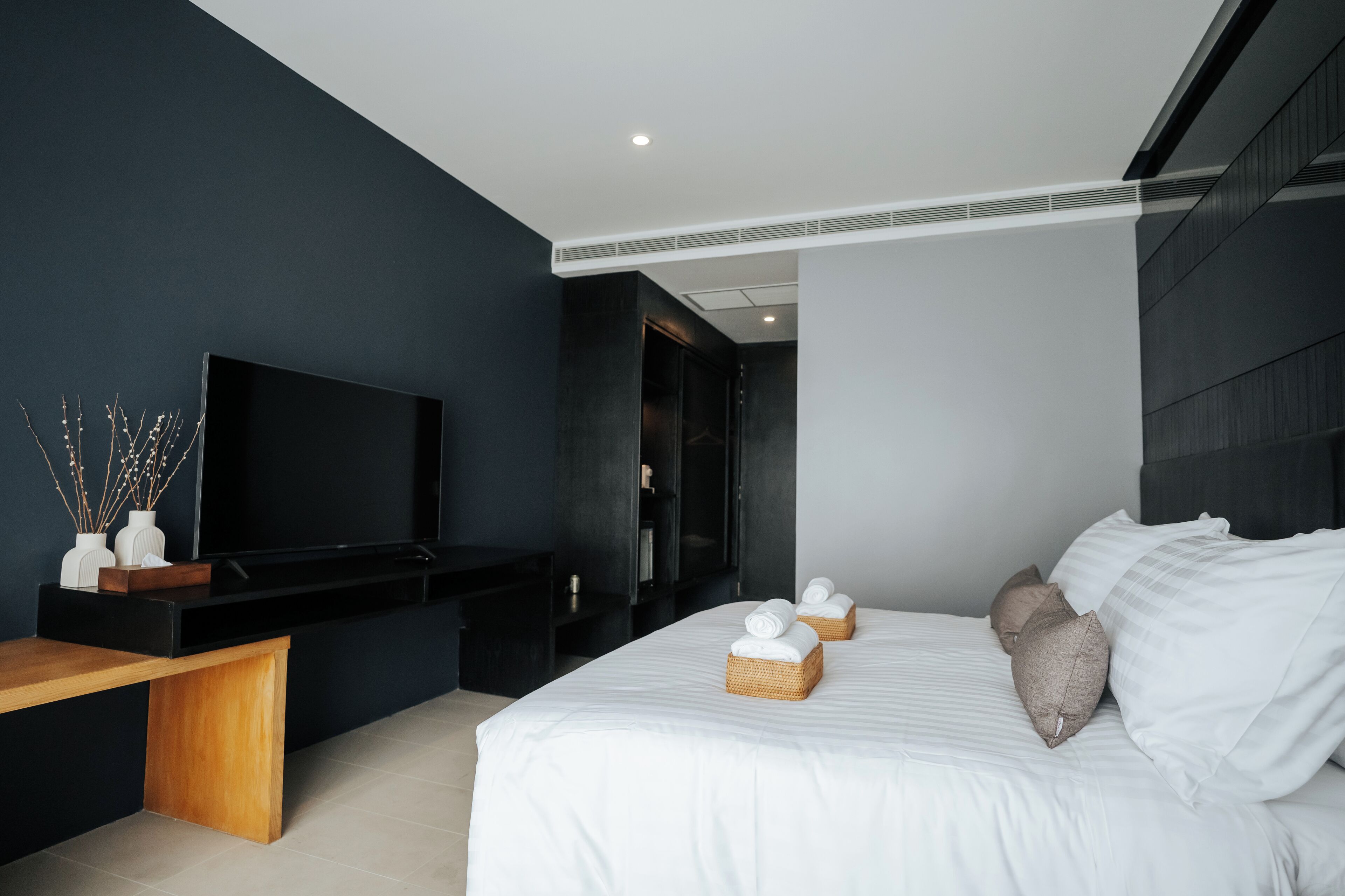 Una habitación de hotel moderna con una cama grande, un televisor de pantalla plana, un banco de madera y un armario de color oscuro.