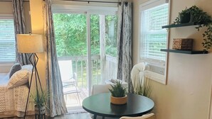 Una habitación acogedora con una mesa redonda, dos sillas, una lámpara de pie y vistas a zonas verdes a través de grandes ventanales.
