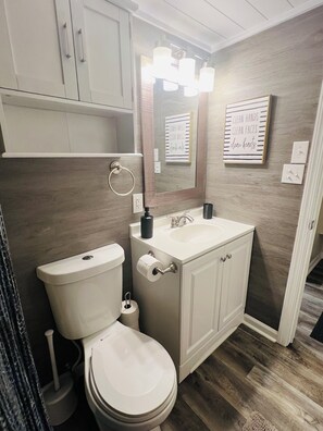 Ein Badezimmer mit Toilette, Waschbecken und Schrank. Über dem Waschbecken befinden sich Leuchten und an der Wand hängt ein gerahmtes Schild.