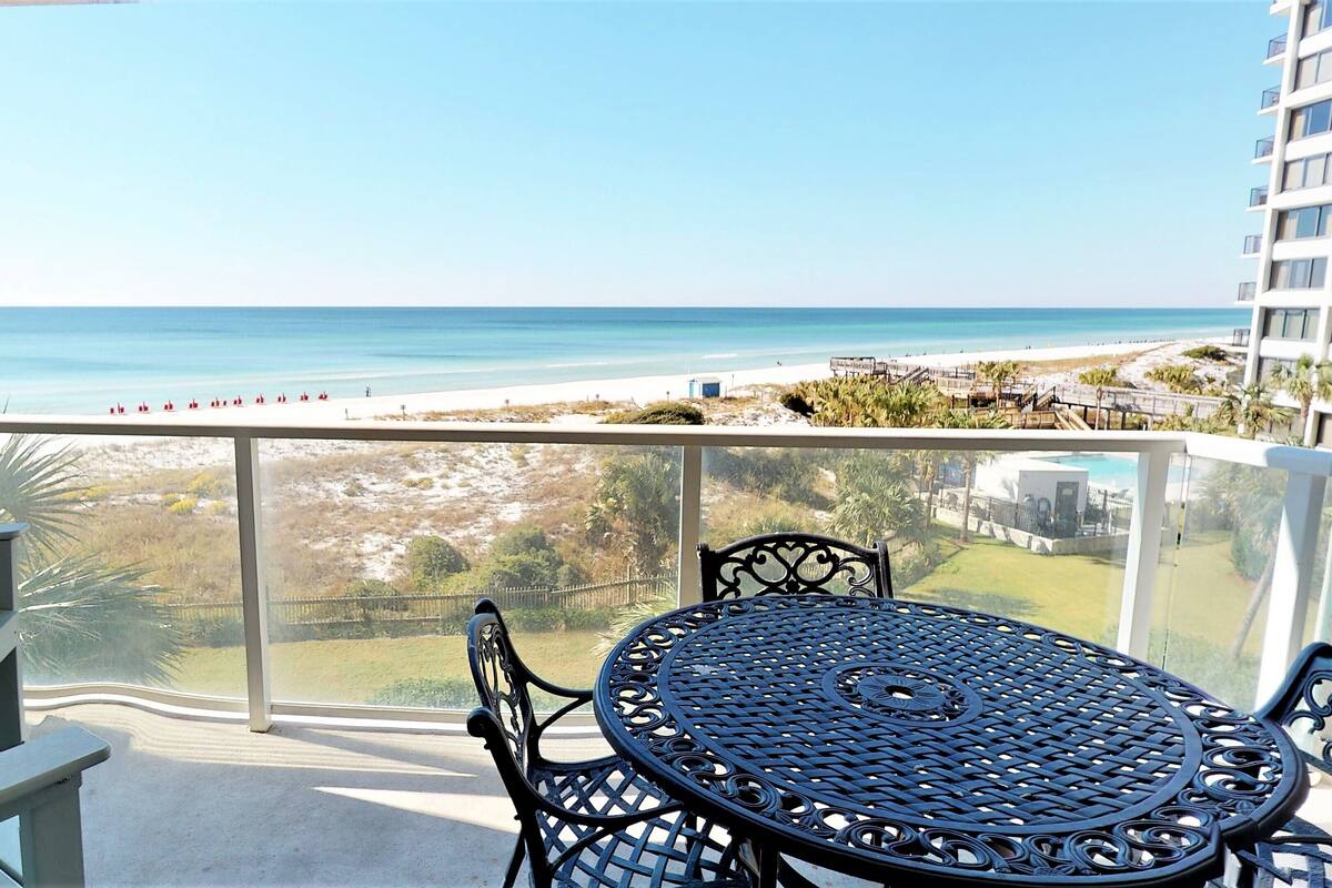 A balcony with a table and chairs, overlooking a beach and ocean.