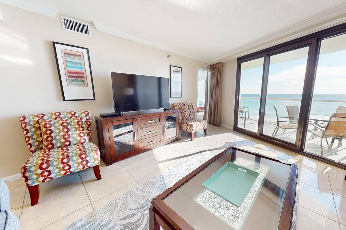 A spacious living room with a large TV, a patterned armchair, a wooden cabinet, and a glass coffee table. There is a balcony with a view of the ocean.