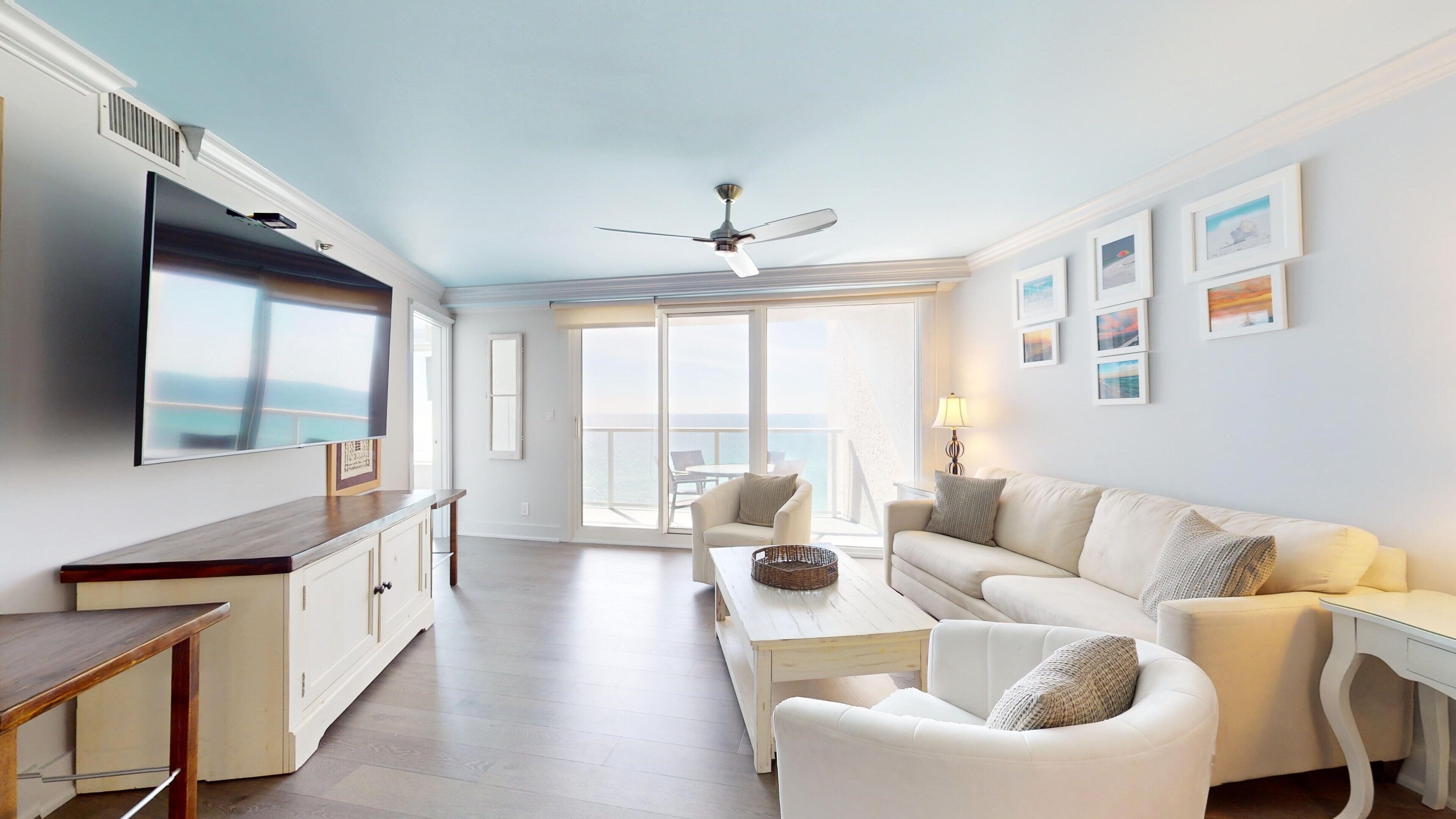 A spacious living area with a large TV, a ceiling fan, a sofa, a coffee table, and a view of the ocean through large windows.