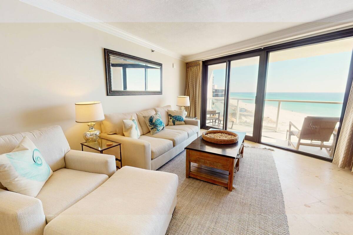 A hotel room with a large window offering a view of the beach, a beige sofa, a small table with a lamp, and a mirror on the wall.