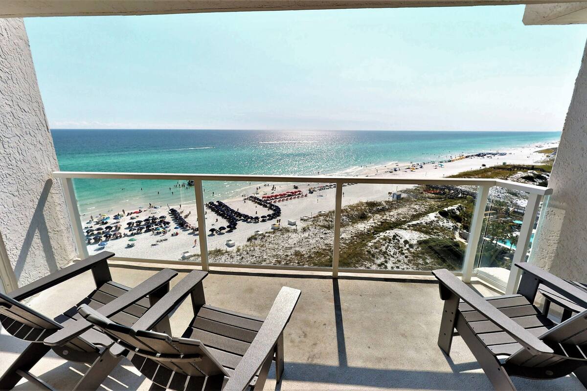 A balcony with two lounge chairs, overlooking a beach with umbrellas and people.
