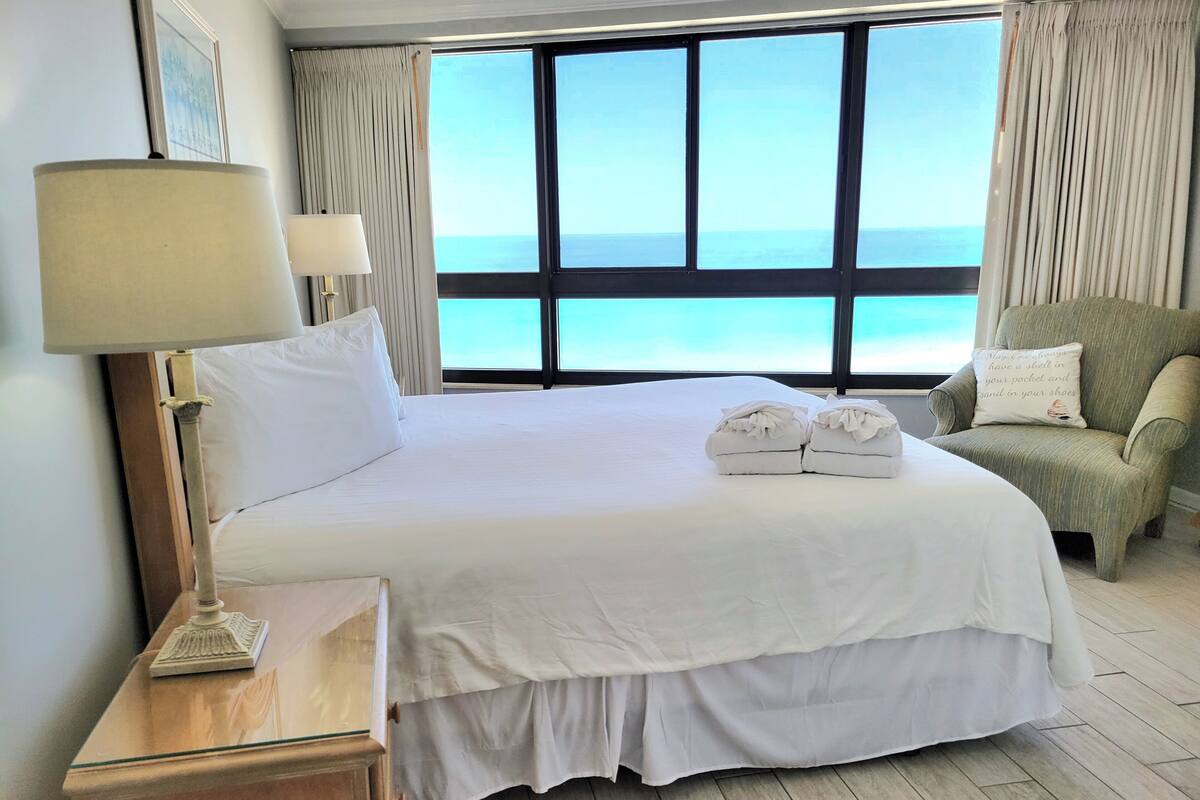 A hotel room with a large bed, a chair, a lamp, and a view of the sea.