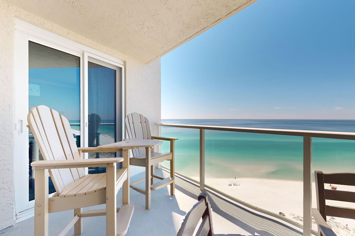A balcony with two Adirondack chairs and a table, overlooking a beach with clear turquoise water.
