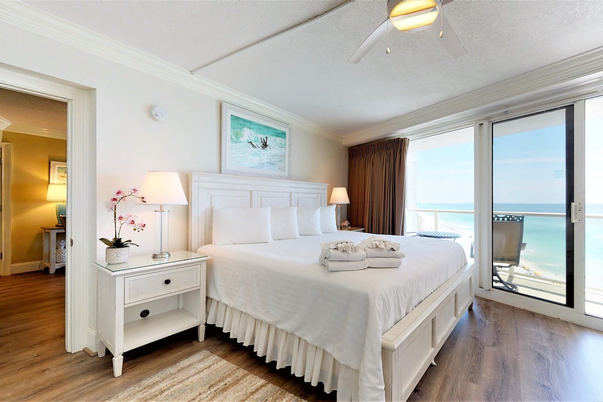 A hotel room with a large bed, two bedside tables with lamps, a view of the ocean through a sliding door, and a ceiling fan.