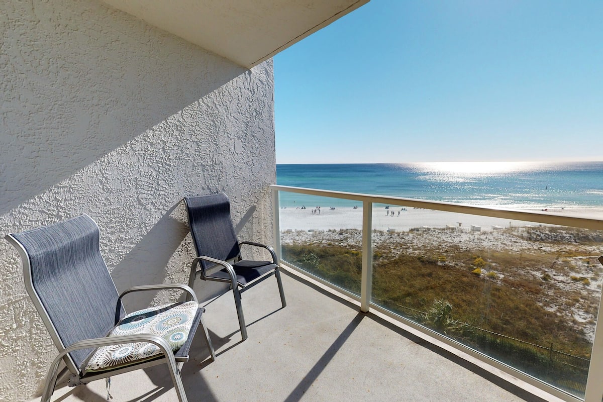 A balcony with two chairs and a table, overlooking a beach with people and a clear sky.