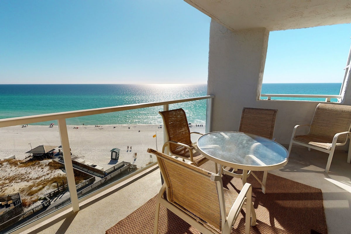 A balcony with wicker chairs and a glass table overlooking a beach with people and a lifeguard tower.