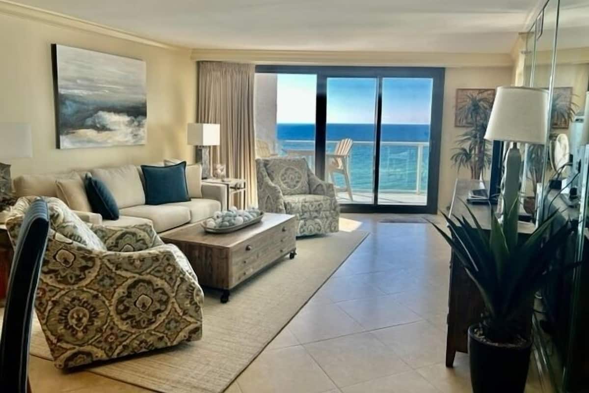A spacious living room with a sofa, armchairs, a coffee table, and a view of the ocean through large sliding doors.