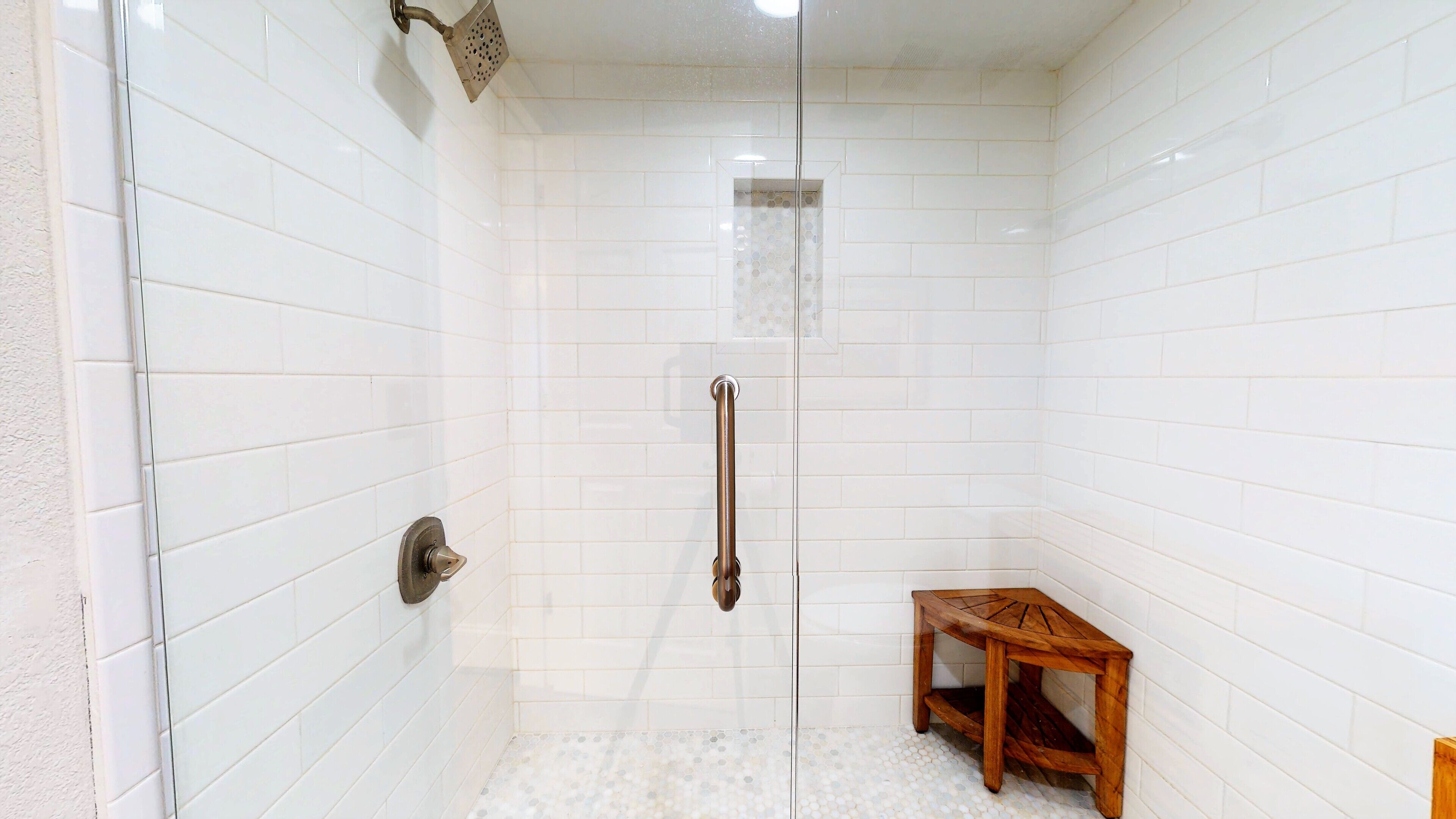 A shower with white tiled walls, a glass door, and a wooden bench.