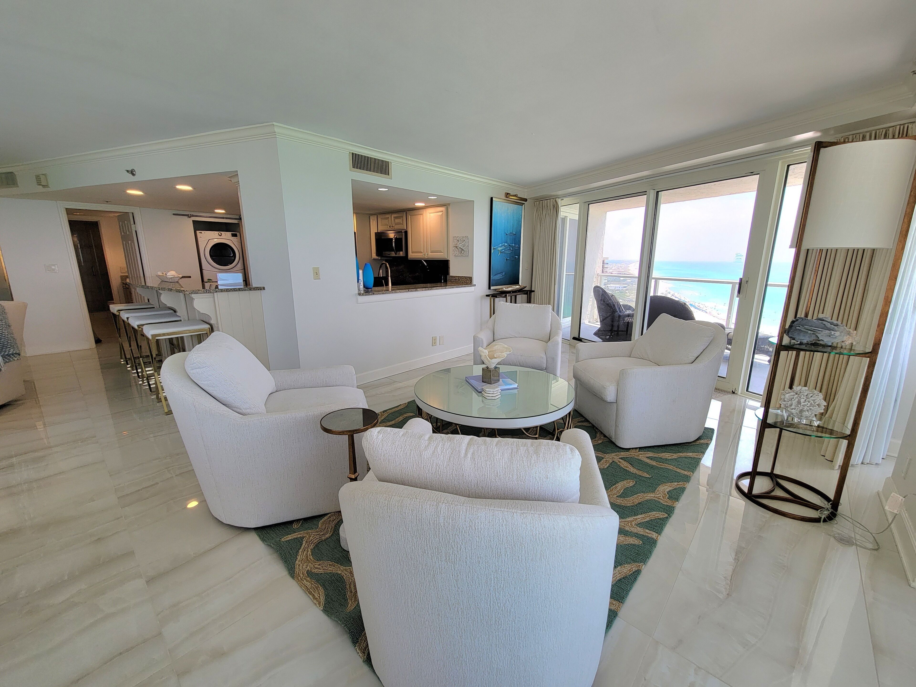 Una sala de estar moderna con una mesa de centro redonda de vidrio, sillones blancos y vistas al océano a través de ventanas amplias.