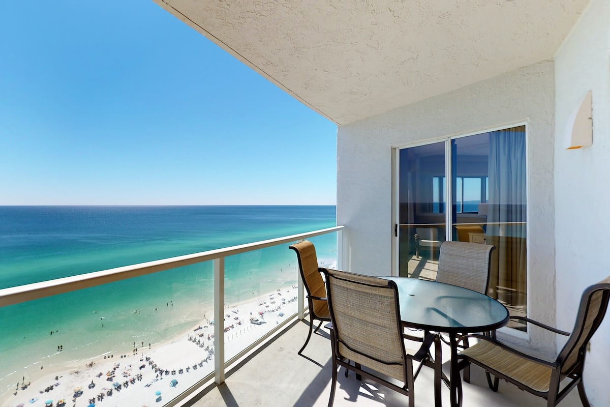 A balcony with a view of a beach and ocean. There is a table and two chairs.