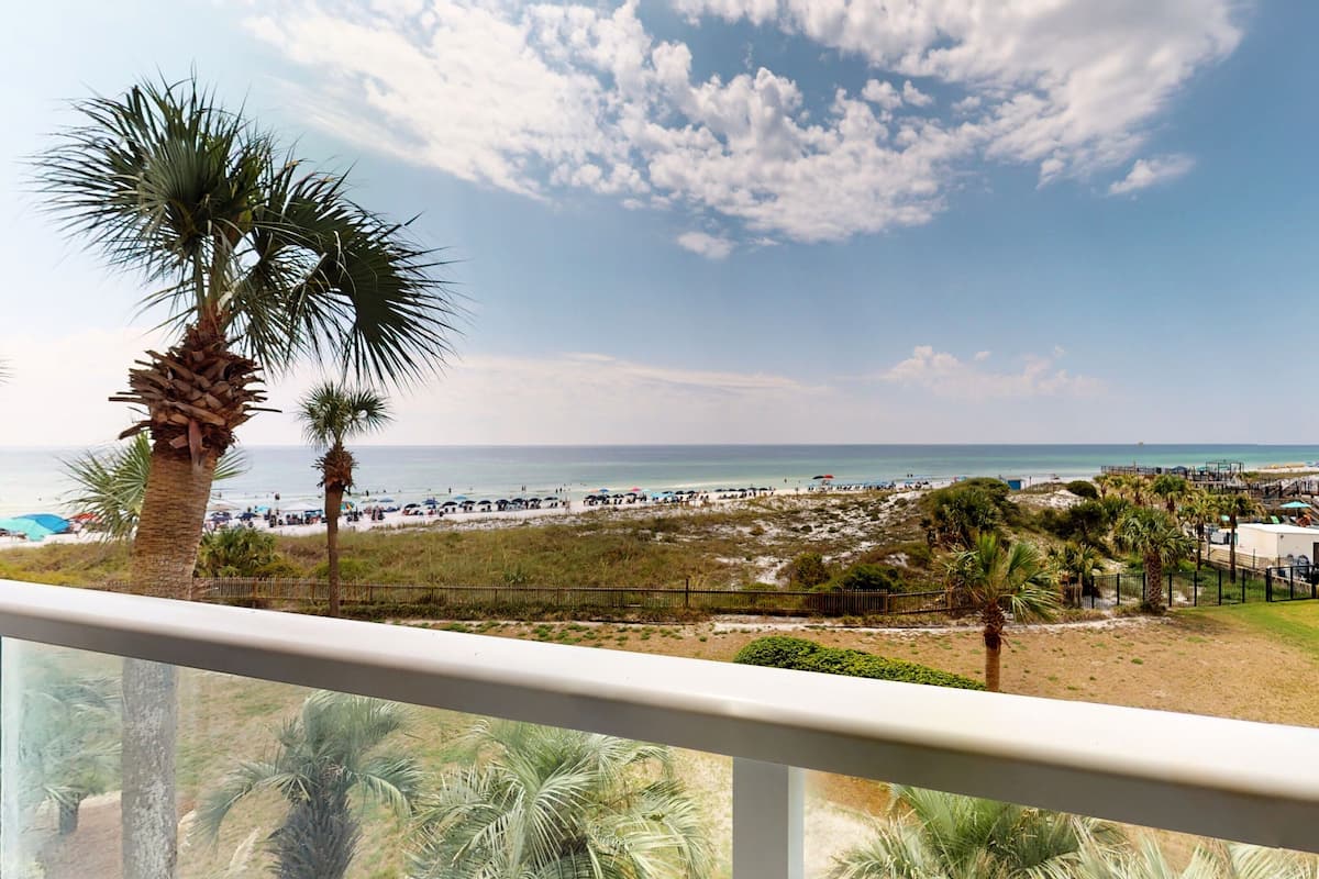 A balcony with a view of a beach, palm trees, and a building with balconies.