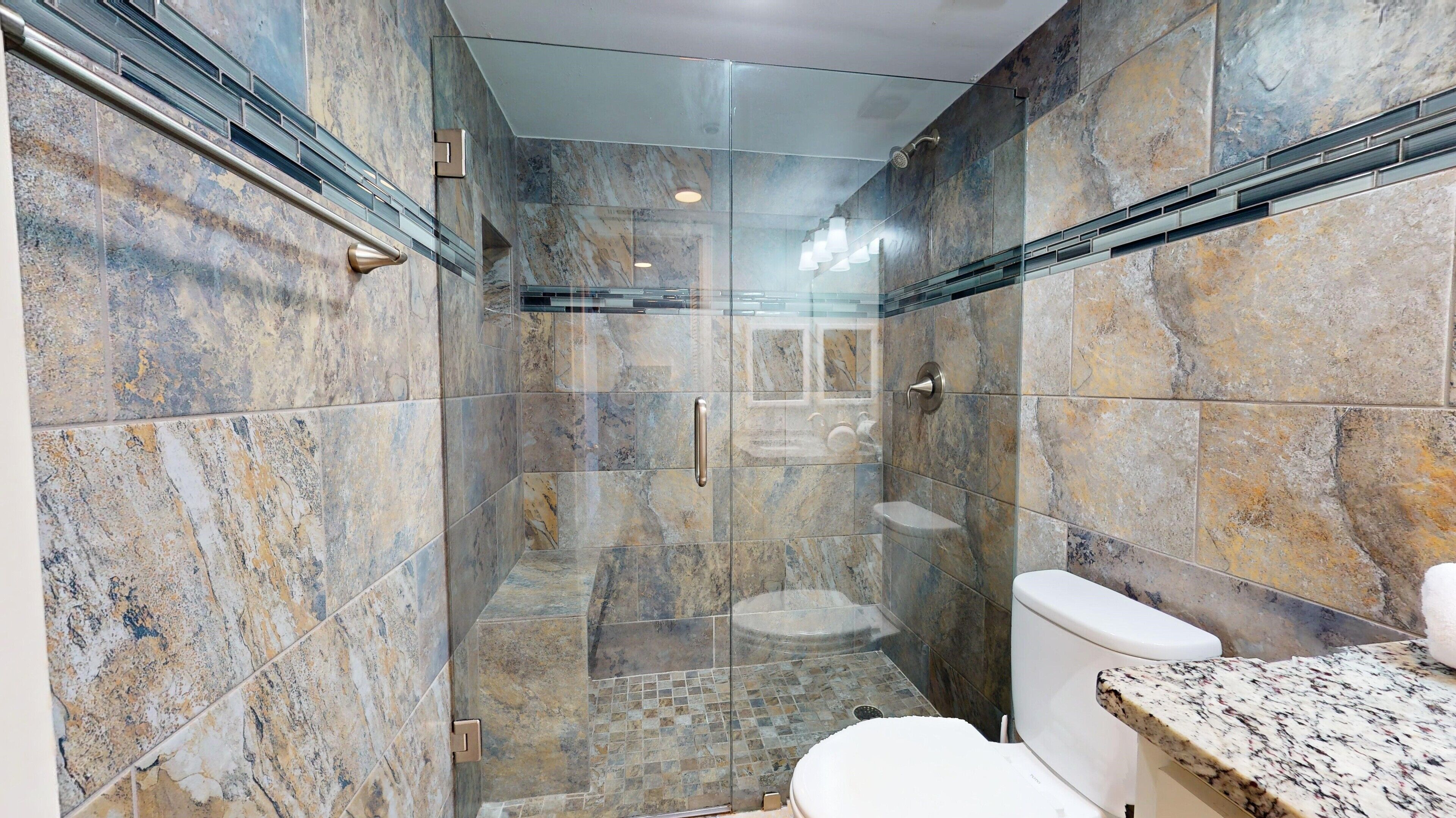 A modern bathroom with a glass-enclosed shower, marble tiles, and a granite countertop.