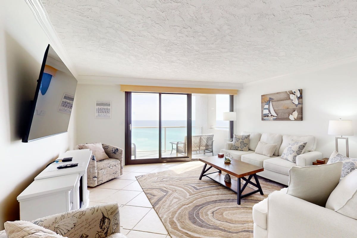 A spacious living room with a large TV, a sofa, a coffee table, and a view of the ocean through the sliding glass doors.
