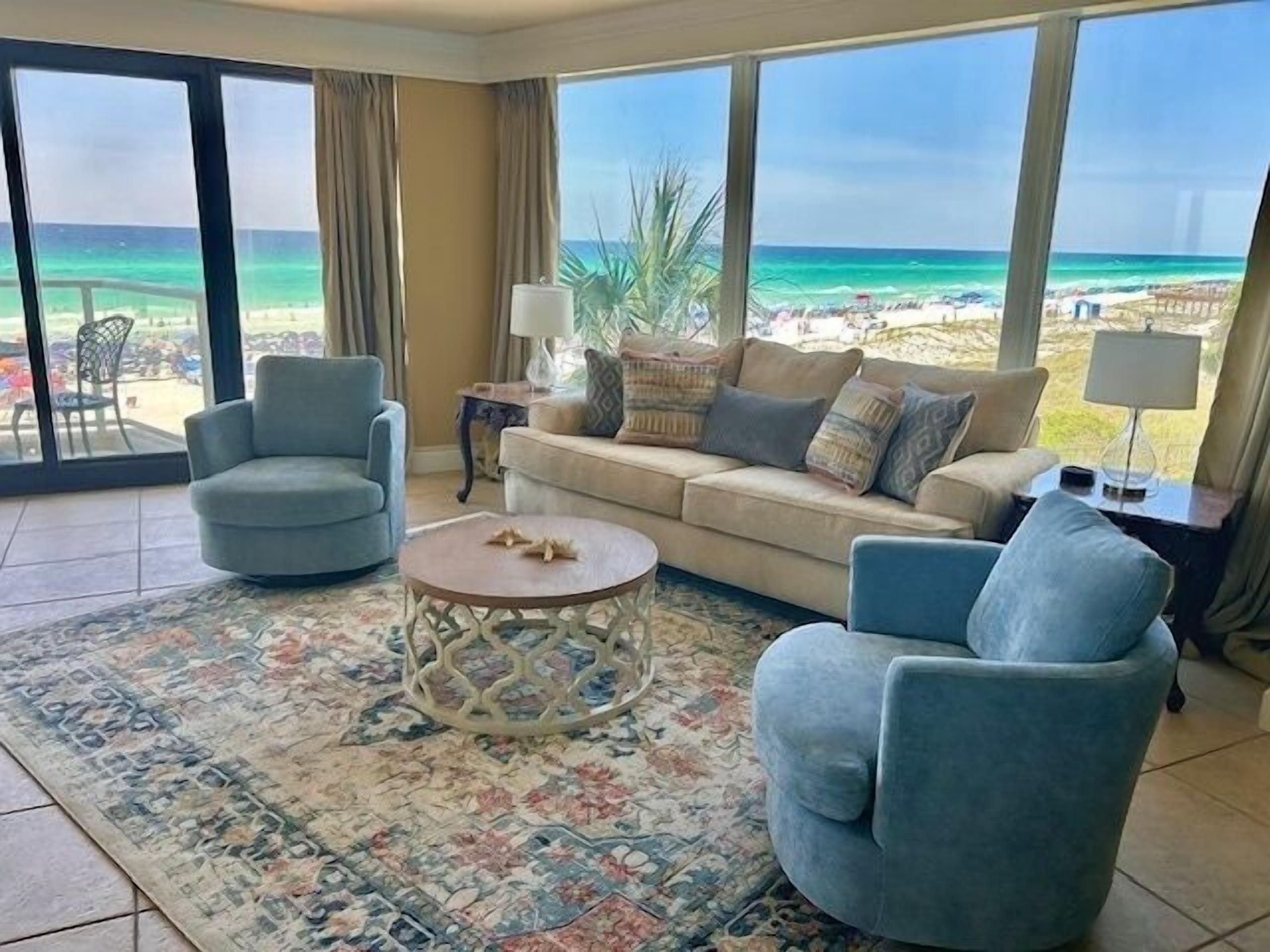 Un salon avec vue sur la plage, comprenant un canapé, deux fauteuils, une table basse et une lampe.