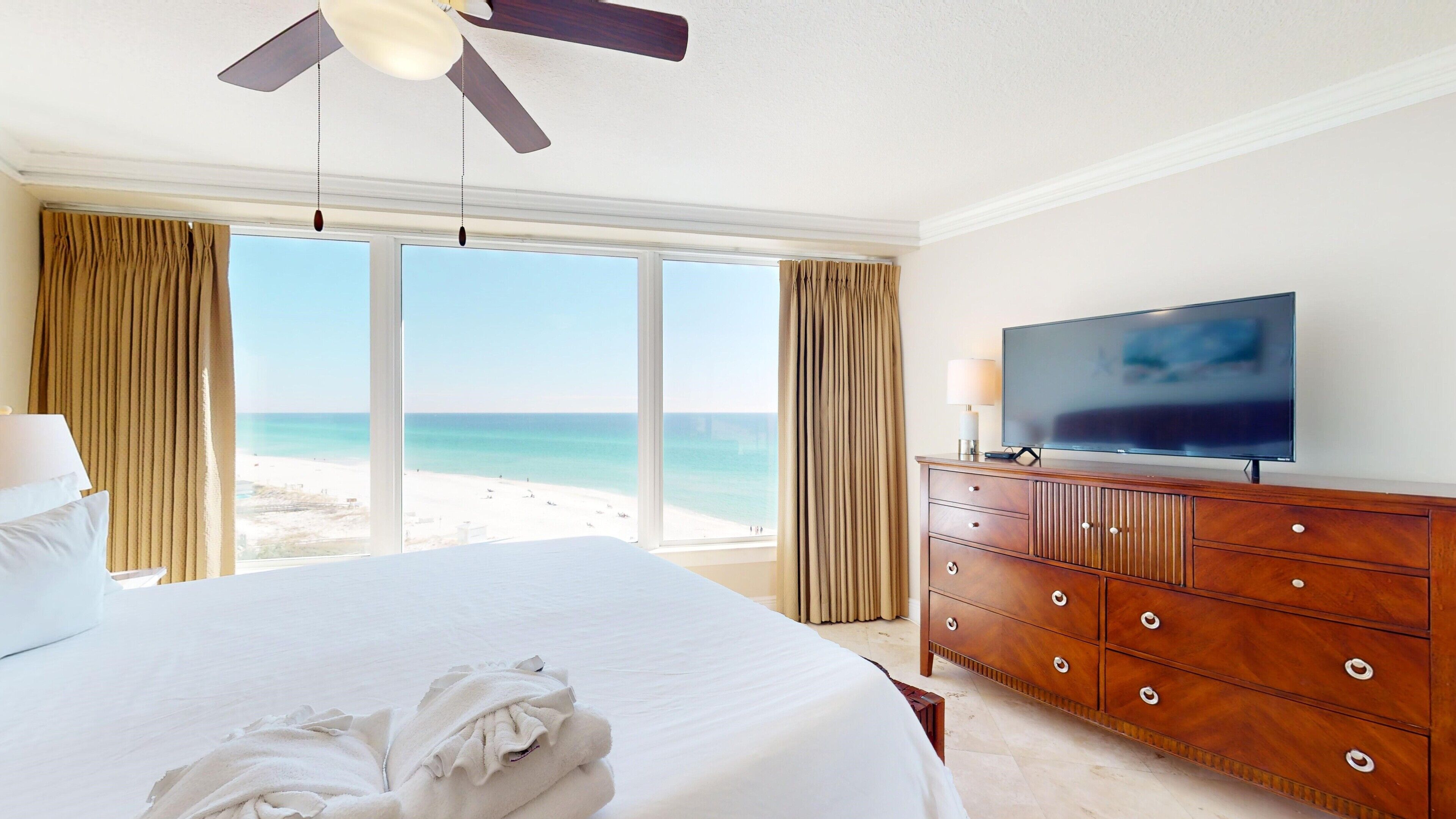 A hotel room with a large bed, a wooden dresser, a flat-screen TV, and a view of the beach.