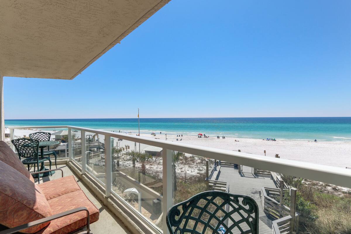 A balcony with a view of a beach, outdoor furniture, and a wooden deck.