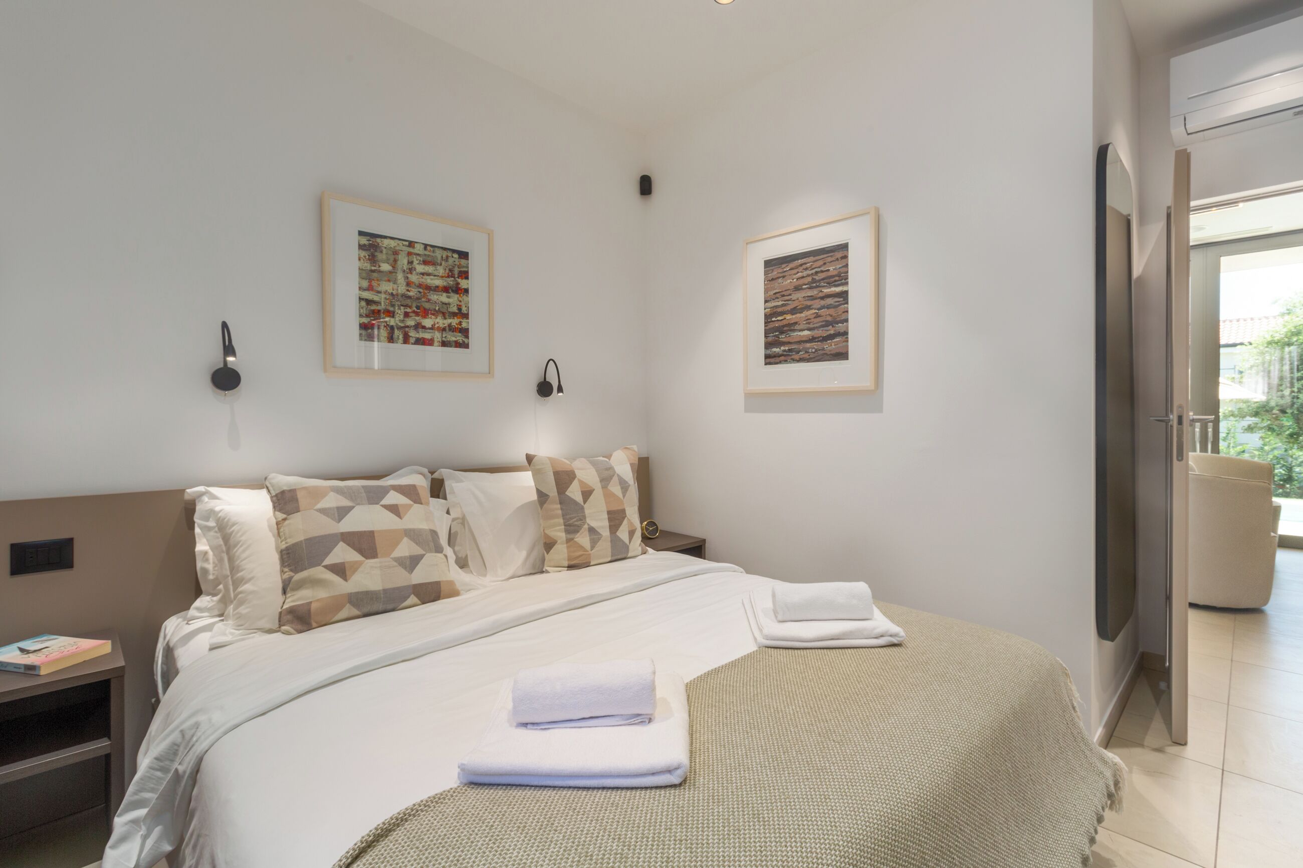A modern hotel room with a neatly made bed, two framed artworks on the wall, a bedside table with a book, and a view of the outdoors through a glass door.