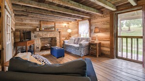 Een rustiek houten interieur met een stenen open haard, houten balken in het plafond, een bank en een groot raam met uitzicht op een tuin.