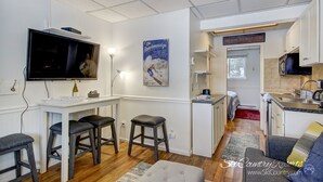 Un espace de vie compact comprenant un coin cuisine, un espace repas avec des tabourets de bar, un téléviseur et une affiche accrochée au mur.