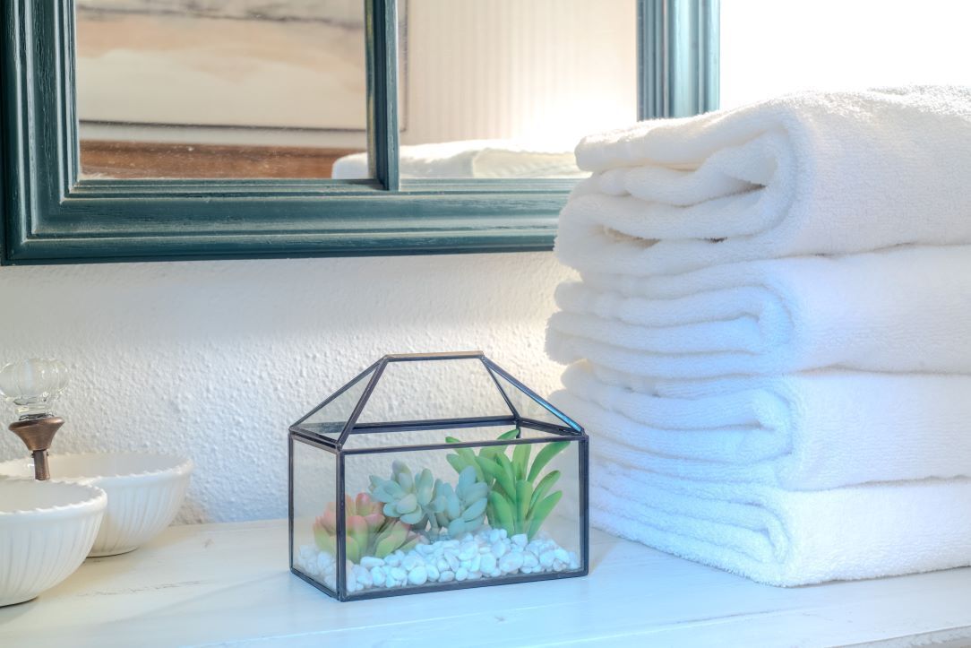 Ein Fenster mit Blick auf einen Strand, ein Stapel weißer Handtücher und ein gläsernes Terrarium mit Sukkulenten und Kieselsteinen.
