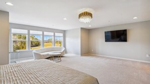 Ein geräumiges Schlafzimmer mit einem großen Bett, einem weißen Sessel, einer Chandelier, einem Flachbildfernseher und großen Fenstern mit Blick ins Freie.