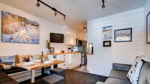 A modern kitchen with white cabinets, a microwave, and a flat-screen TV. There is a dining area with a wooden table and benches.
