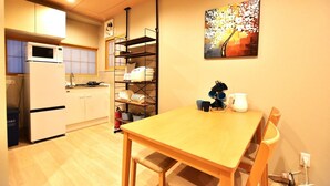 A compact kitchen with a microwave, stove, and shelves with dishes. A dining table with chairs is in the foreground, and a painting hangs on the wall. - Tamatsukan (Osaka)