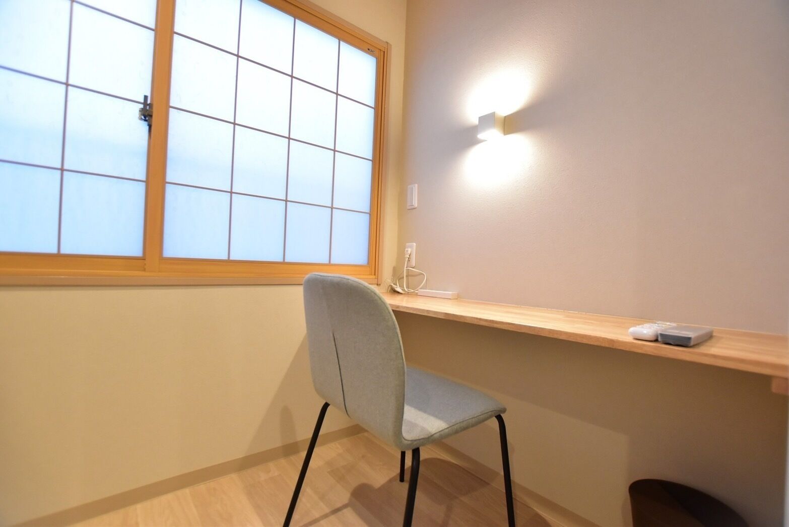 Una habitación con ventana, una silla, una estantería con un control remoto y una lámpara de pared.