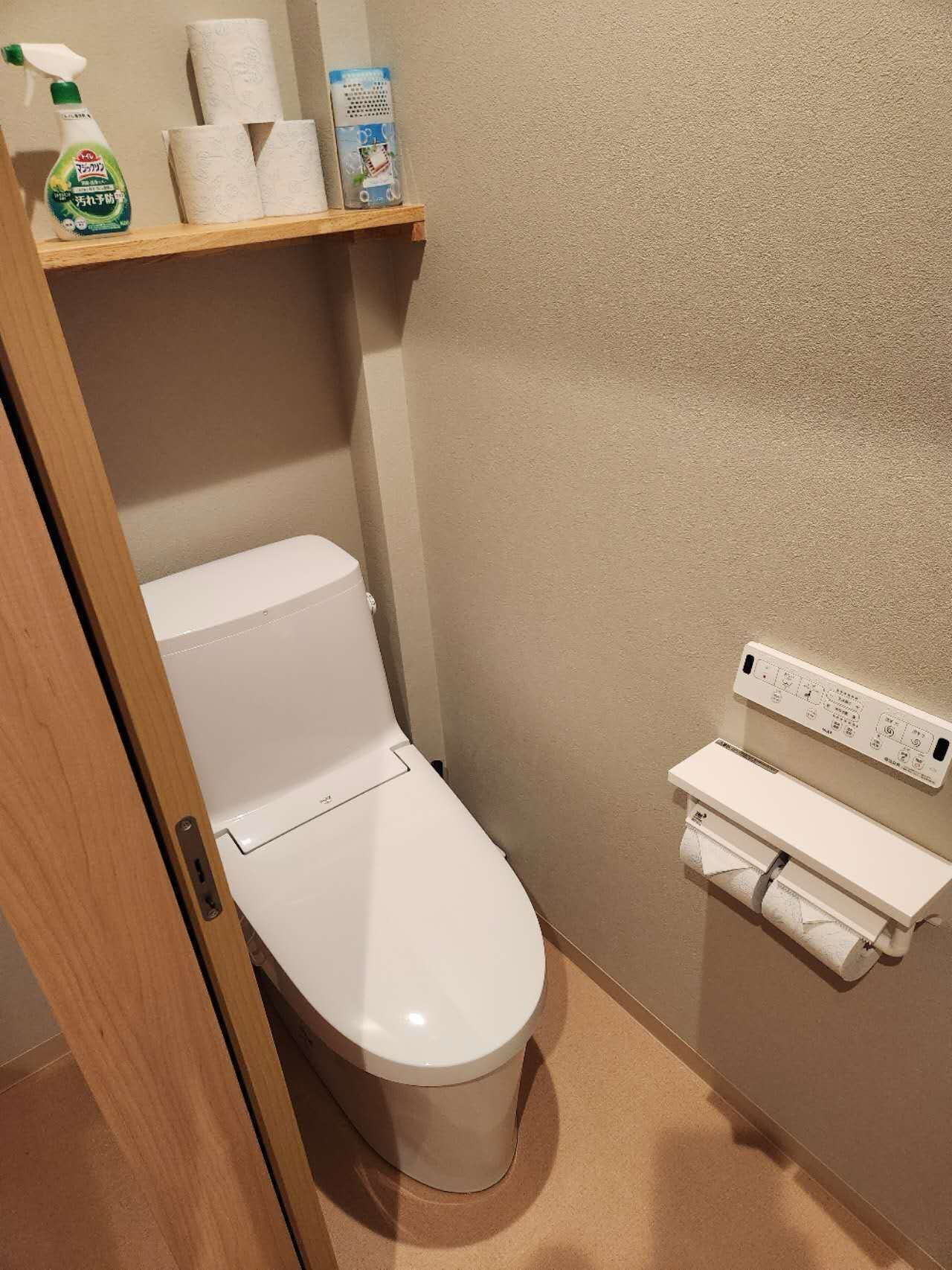 Ett litet badrum med en vit toalett, en hylla med toalettpapper och rengöringsmedel samt en väggmonterad toalettpappershållare.