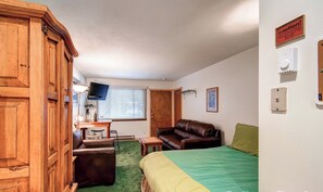 A compact room with a bed, a brown leather sofa, a small wooden table, a television mounted on the wall, and a window with blinds. - Budget-Friendly Top Floor Unit in Downtown - Walk to Shopping/Dining | PM7B (Breckenridge)