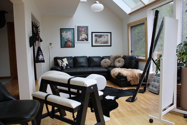 A modern living room with a black leather sofa, a standing exercise bike, and a large window providing natural light.