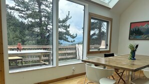 A modern dining area with a wooden table and white chairs, large windows offering a view of a deck and trees, and a painting on the wall.