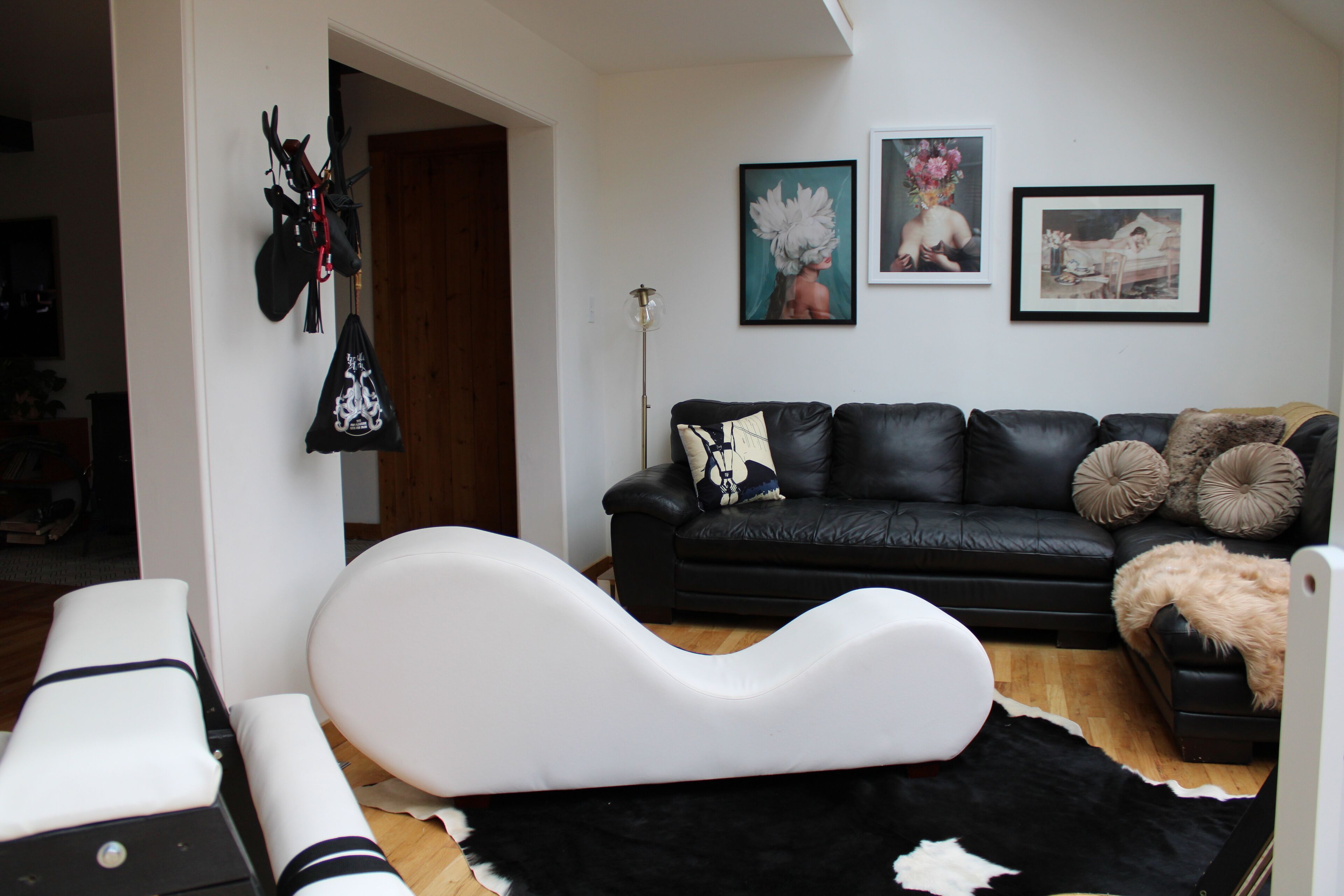A modern living room with a black leather sofa, white accent pillows, and a black and white rug. There are two framed artworks on the wall and a black bag hanging on the wall.