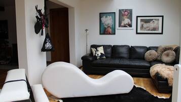 A modern living room with a black leather sofa, white accent pillows, and a black and white rug. There are two framed artworks on the wall and a black bag hanging on the wall.