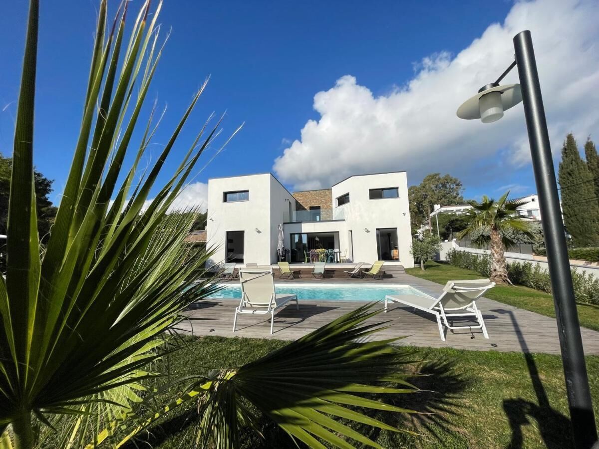 A modern two-story building with a swimming pool, surrounded by greenery and a clear sky.