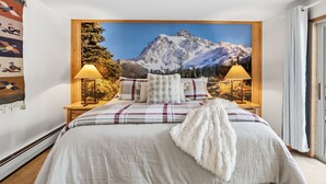 Ein Schlafzimmer mit Blick auf die Berge, ein Bett mit kariertem Überwurf, zwei Nachttischlampen und ein Holz-Kopfteil.