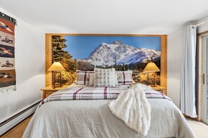 A bedroom with a mountain view, a bed with a plaid bedspread, two bedside lamps, and a wooden headboard.