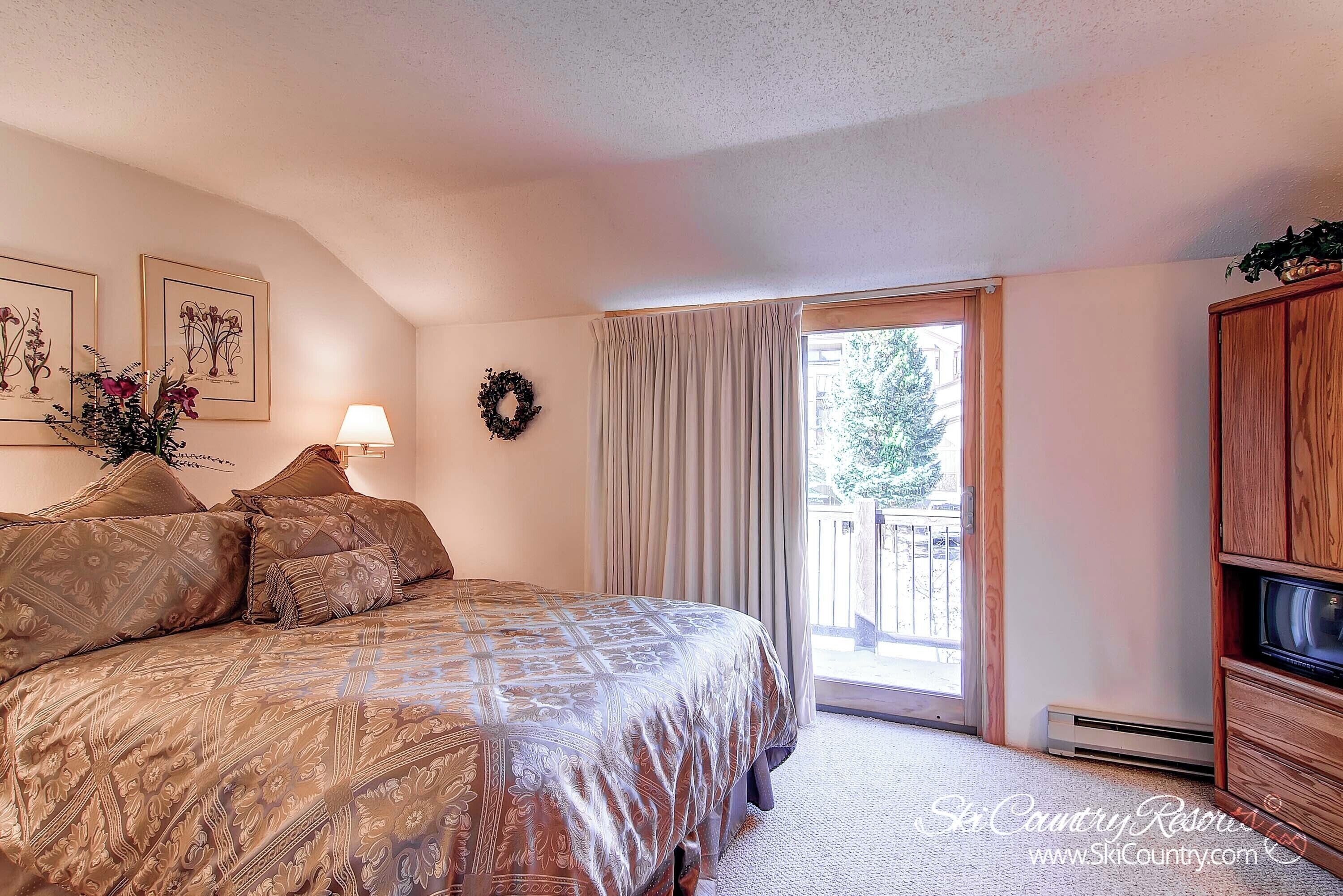 A bedroom with a large bed, a window with curtains, a wall-mounted wreath, and a wooden cabinet.