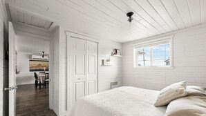 Un dormitorio con una cama grande, paredes blancas, suelo de madera, una ventana y una puerta que da a otra habitación.