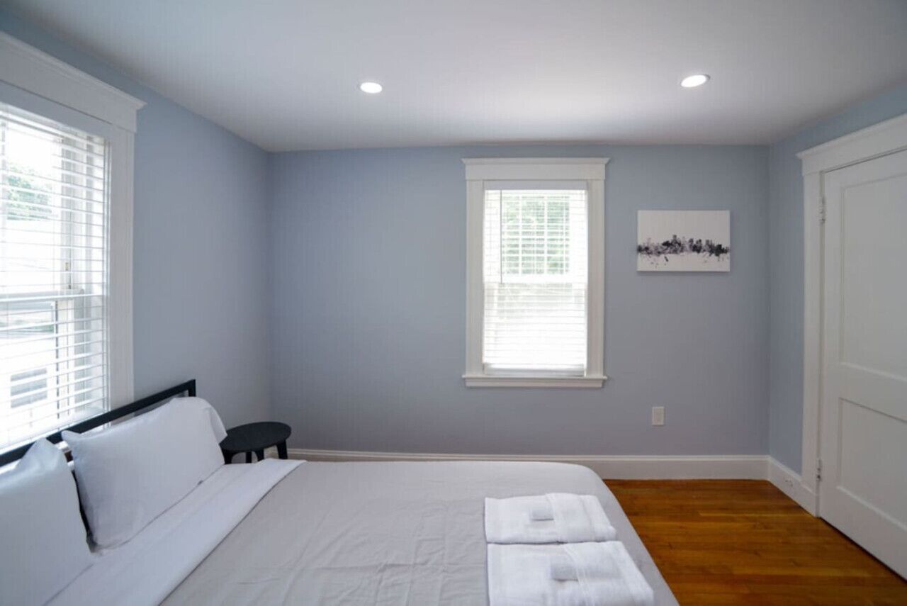 A bedroom with a bed, two windows, a small table, and a painting on the wall.
