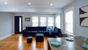 Uma sala de estar moderna com um sofá seccional escuro, uma pequena mesa de centro redonda e uma mesa de jantar posta com pratos e copos.